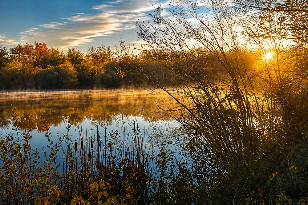 Sun rising over a lake with mist rising from the water, surrounded by wildernessand sunlight shining through the trees - Pocasset Pokanoket Land Trust