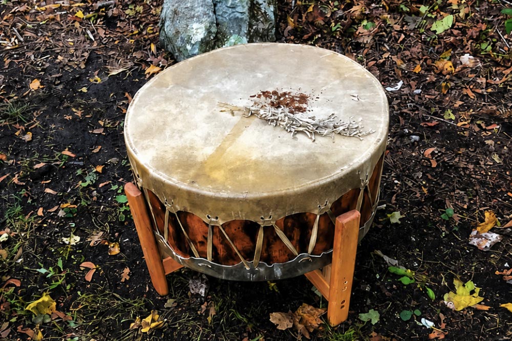 Hide drum on a wooden stand outdoors, with a sage bundle and loose herbs resting on the drumhead.