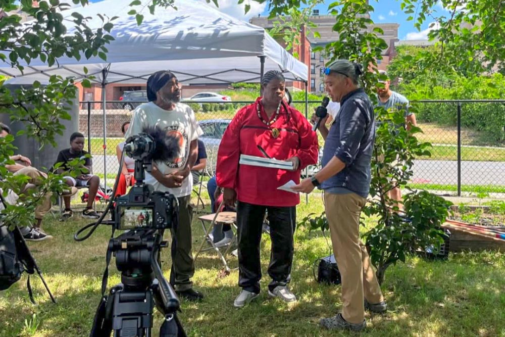 Two people being interviewed on camera at an outdoor community garden event, with a pop-up tent behind them.