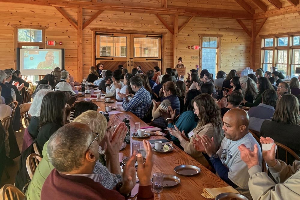 Attendees applaud at long wooden tables during a presentation in a timber-frame hall.