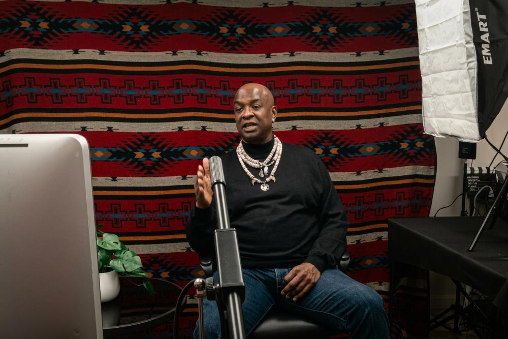 Person speaking into a microphone during a podcast recording, seated in front of a patterned blanket backdrop.