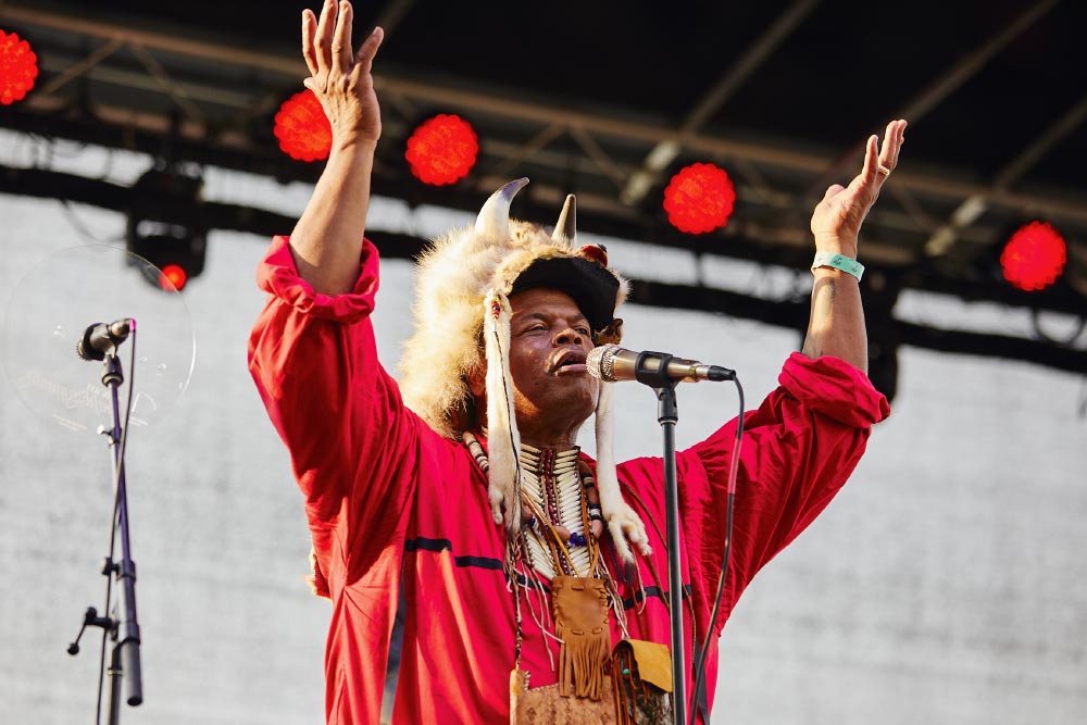 Tribal elder in regalia raises both hands at a microphone while offering a blessing on an outdoor festival stage.